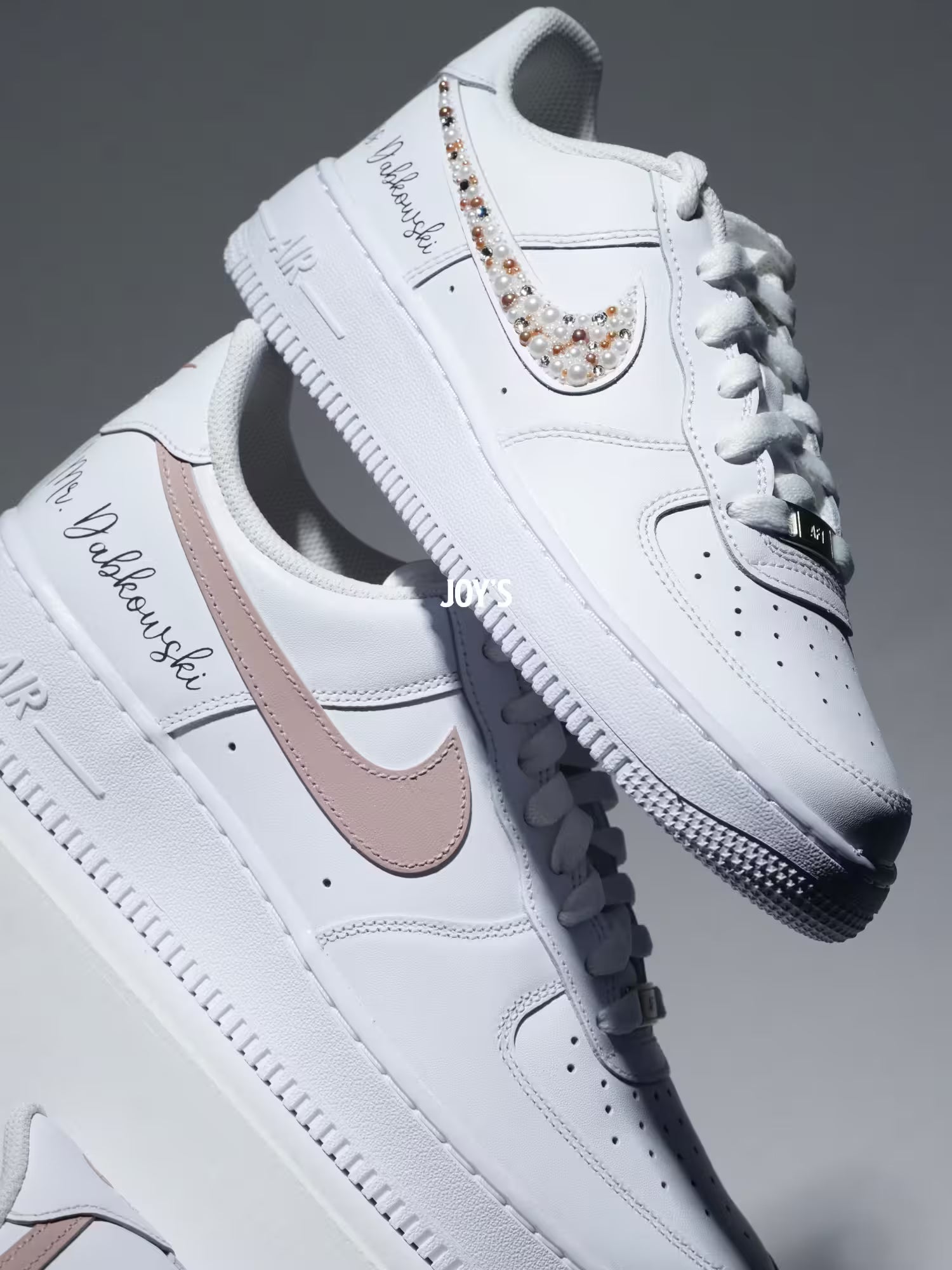 Custom white Nike Air Force 1 sneakers with subtle script lettering, one blush pink swoosh, and one swoosh embellished with pearls and rhinestones.