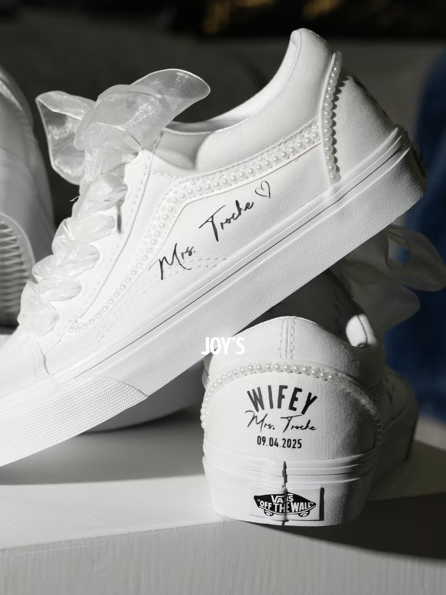 White custom wedding sneakers with pearl trim and personalized bride and groom names, wedding date, and “Wifey” heel text shown in a close-up detail view