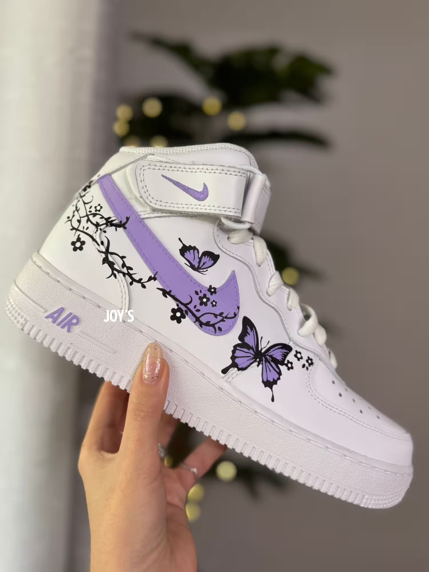 White Nike Air Force 1 Mid sneaker with standard flat white laces, ankle strap, and a lavender swoosh decorated with black floral vines and purple butterfly artwork, held in hand against a softly blurred indoor background.