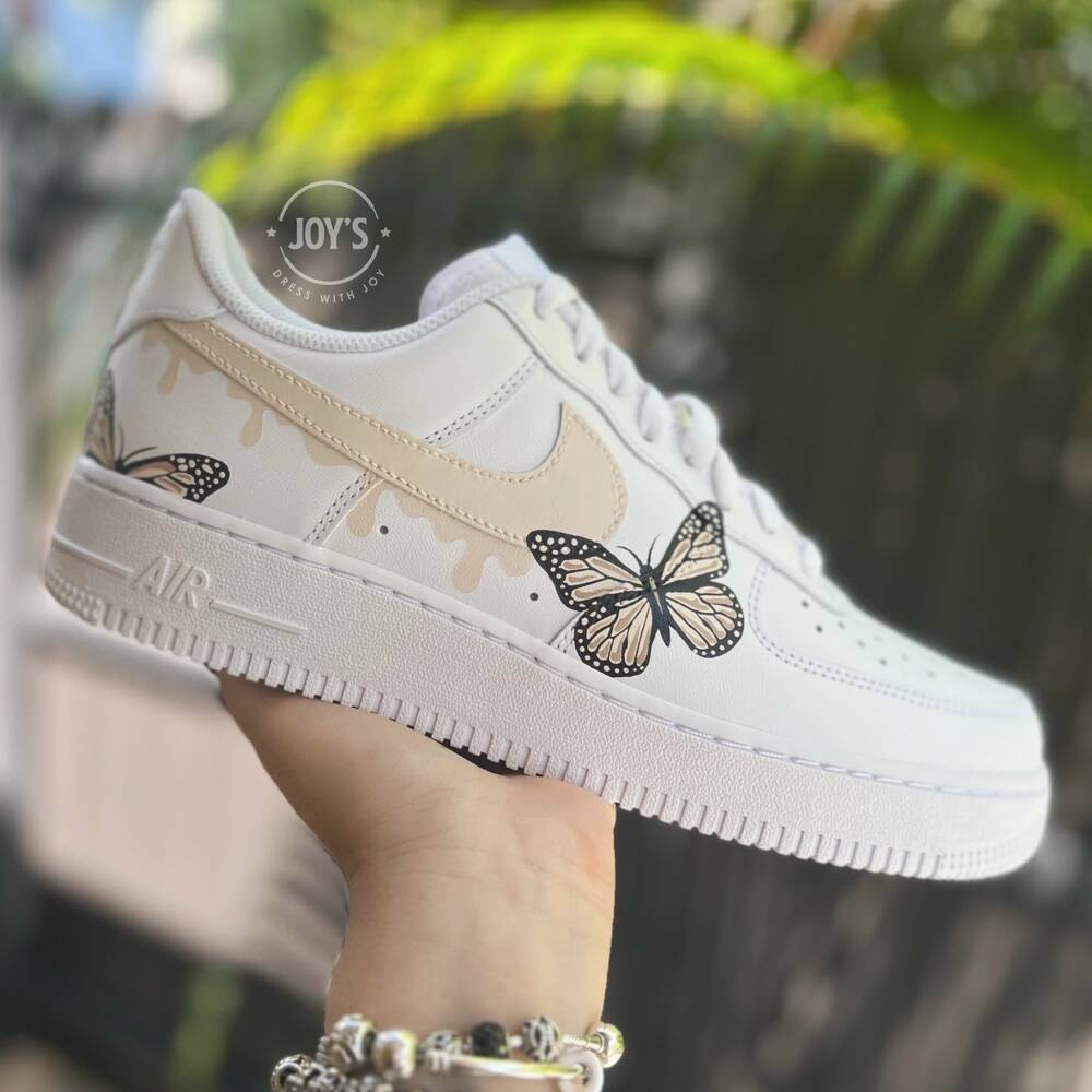 White custom Nike Air Force 1 sneakers with hand-painted butterfly artwork and soft beige drip accents, shown in a hand-held outdoor side view