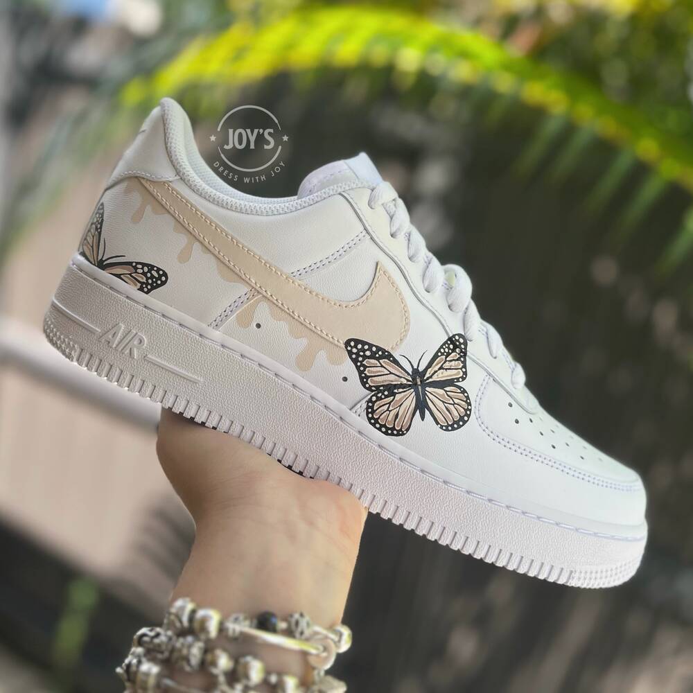 White custom Nike Air Force 1 sneakers with hand-painted butterfly artwork and soft beige drip accents, shown in a hand-held outdoor side view