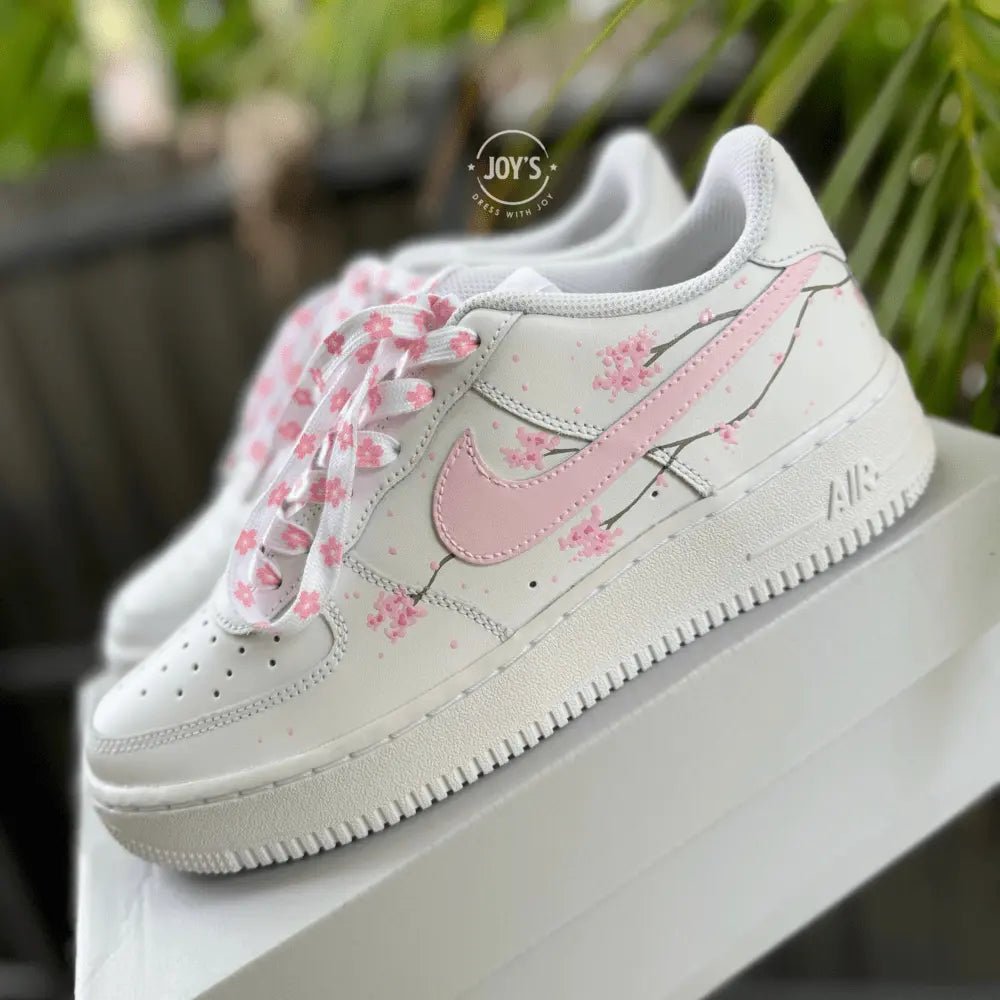 White Nike Air Force 1 with soft pink swoosh and hand-painted cherry blossom branches, shown in close-up.