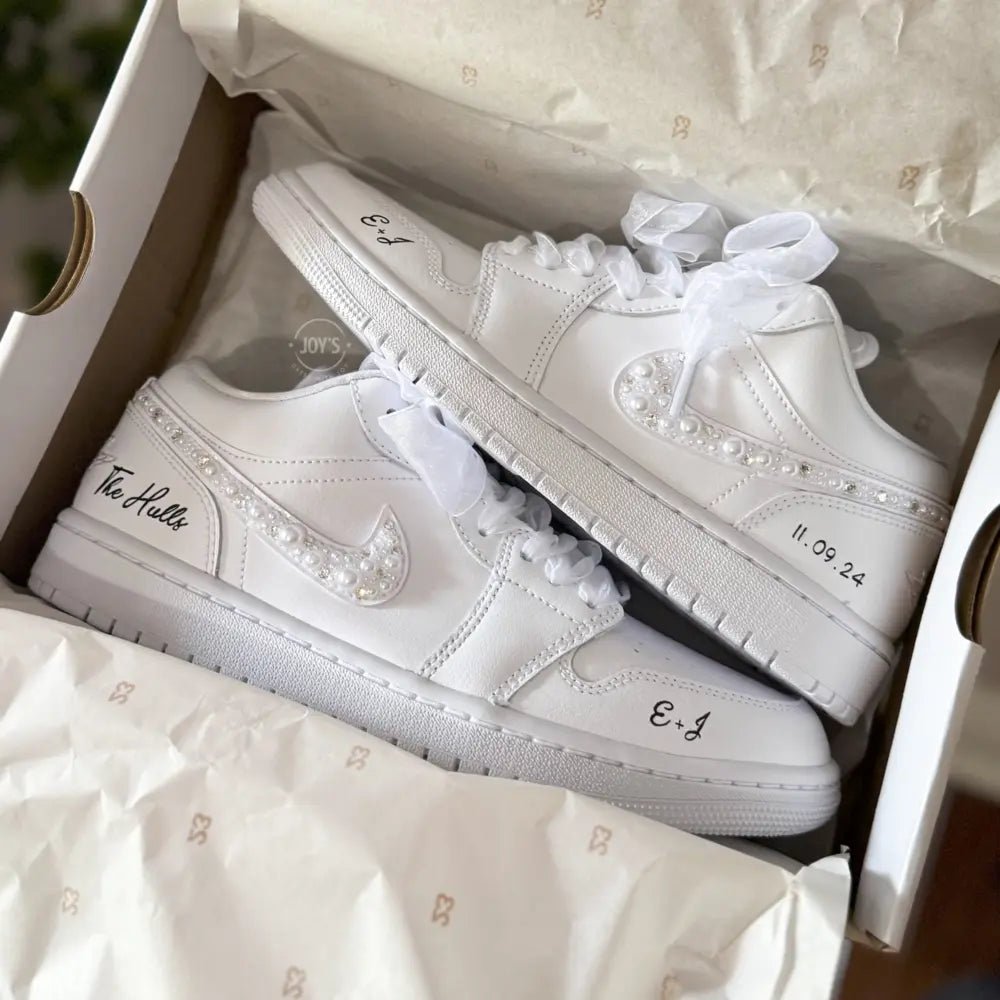 White Nike Air Force 1 Low sneakers (standard sole, not platform), customized with a crystal-embellished swoosh and subtle rhinestone detailing along the panels. Finished with sheer white ribbon laces and personalized black script on the heel and toe, giving a clean, elegant bridal look.