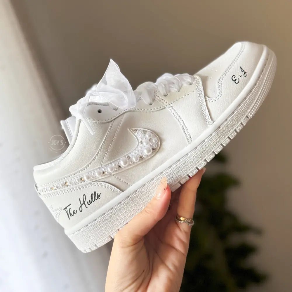 White Nike Air Force 1 Low sneakers (standard sole, not platform), customized with a crystal-embellished swoosh and subtle rhinestone detailing along the panels. Finished with sheer white ribbon laces and personalized black script on the heel and toe, giving a clean, elegant bridal look.
