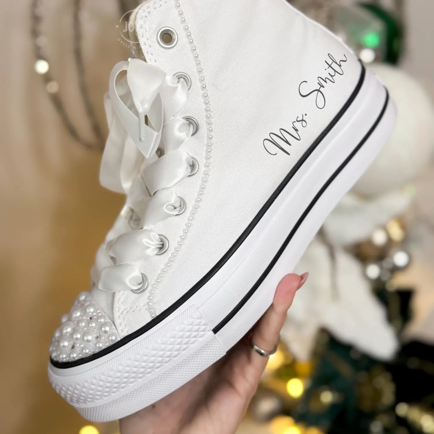 White platform Converse Chuck Taylor wedding sneakers with pearl-embellished toe cap and satin ribbon laces, shown in a hand-held close-up view
