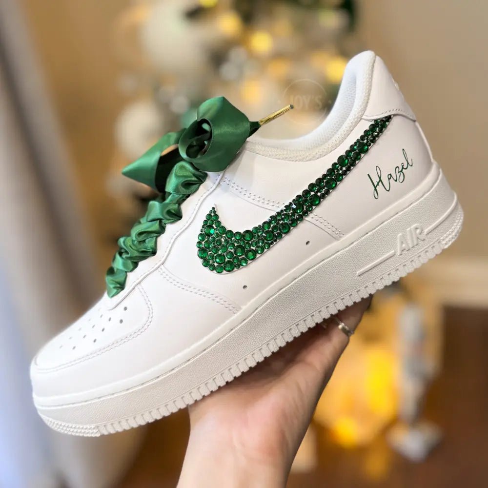 White custom Nike Air Force 1 sneakers with emerald green rhinestone swoosh, satin ribbon laces, and personalized name printed on the side, shown in a hand-held side view