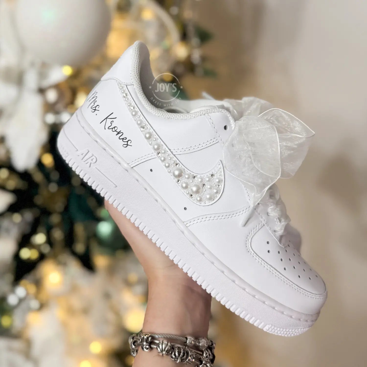 Custom wedding white Nike Air Force 1 with pearl and rhinestone swoosh, white ribbon laces, and the name Mrs. personalized on the heel, shown held in hand with soft holiday lights in the background.