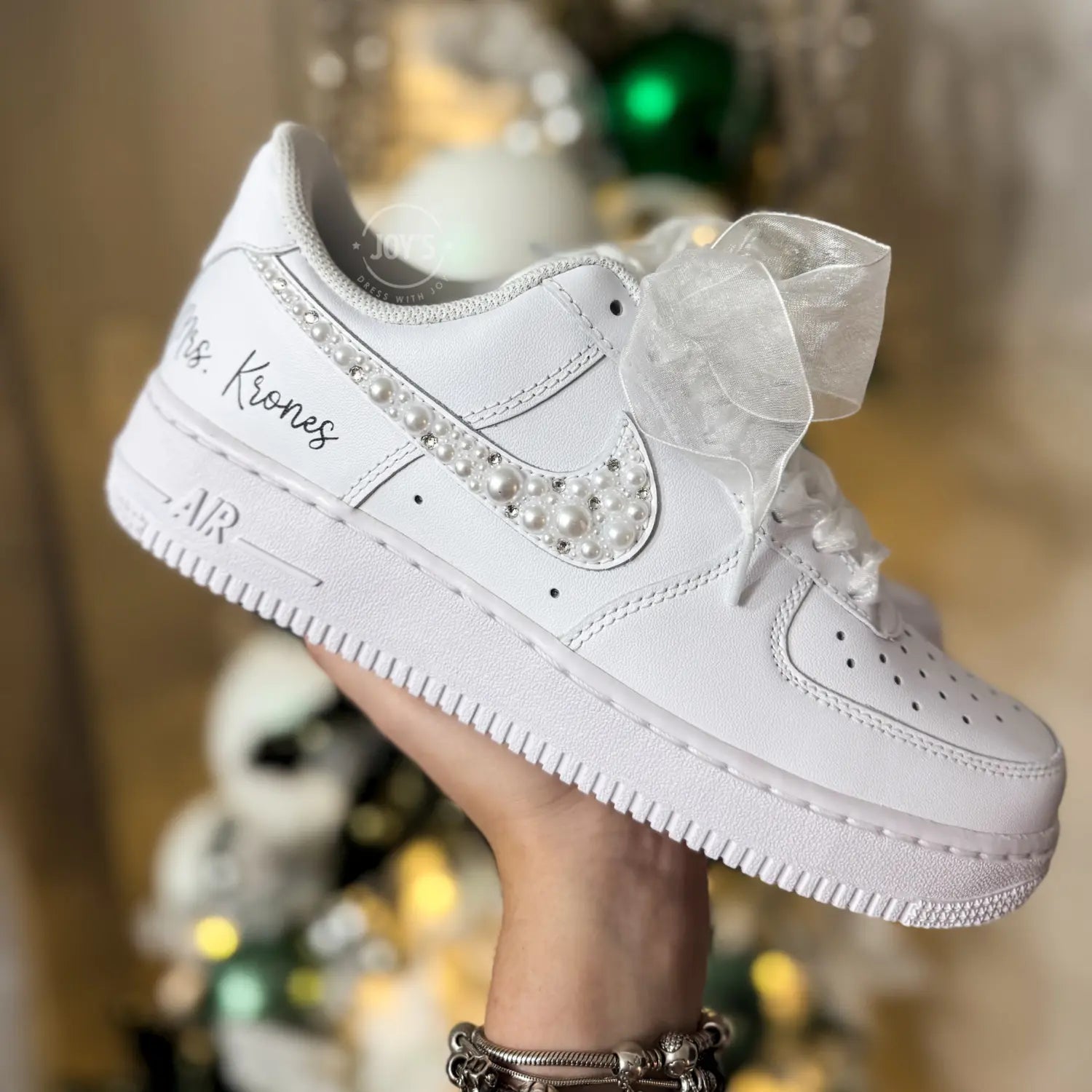 Custom wedding white Nike Air Force 1 with pearl and rhinestone swoosh, white ribbon laces, and the name Mrs. personalized on the heel, shown held in hand with soft holiday lights in the background.