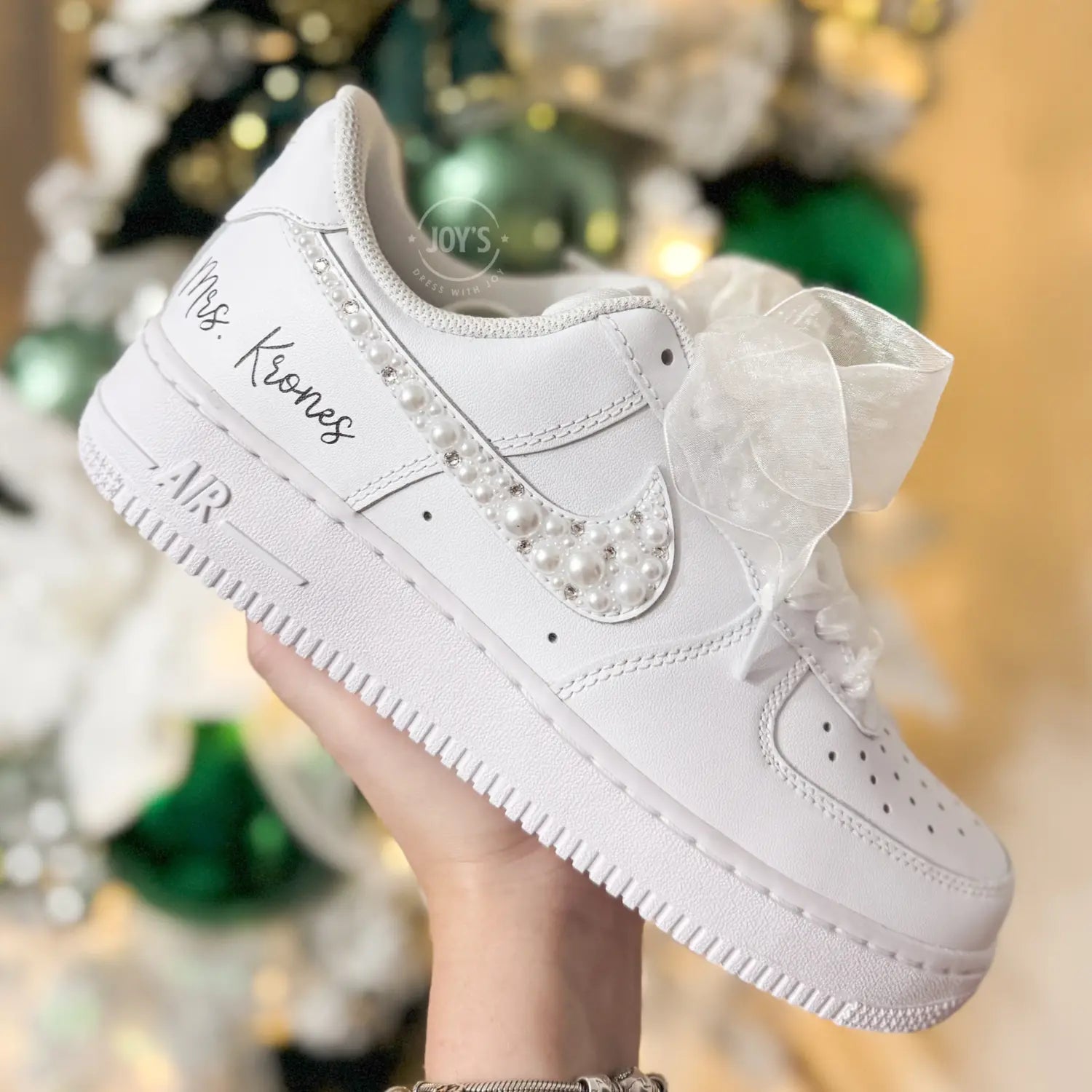 Custom wedding white Nike Air Force 1 with pearl and rhinestone swoosh, white ribbon laces, and the name Mrs. personalized on the heel, shown held in hand with soft holiday lights in the background.
