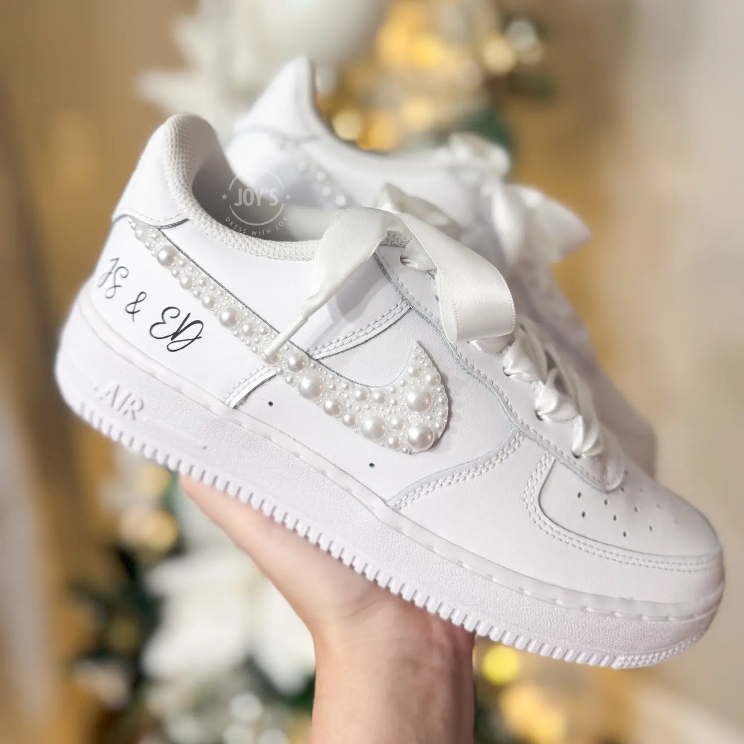 Custom white wedding Nike Air Force 1 with pearl-embellished swoosh, white ribbon laces, and personalized initials “J & E” on the heel, shown held in hand with soft bokeh lights in the background.