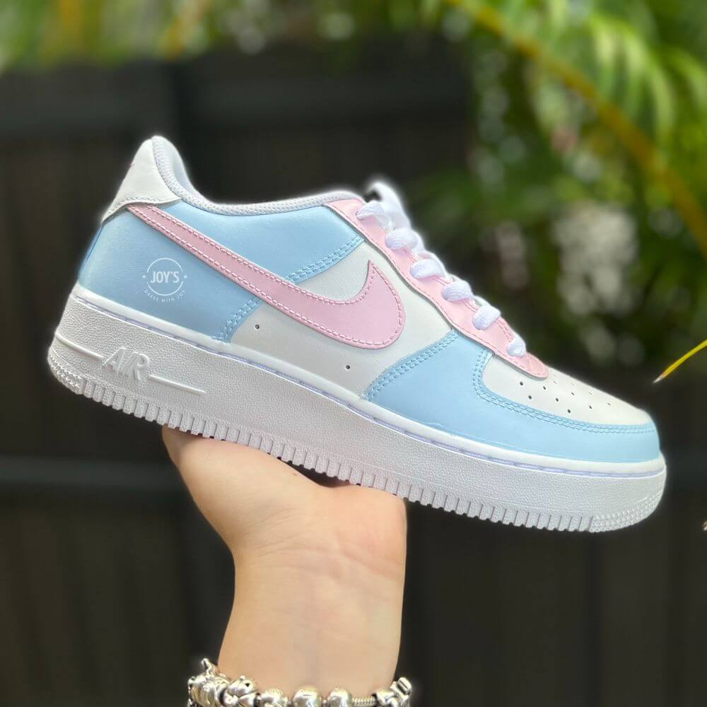White Nike Air Force 1 sneakers with baby blue panels, soft pink swoosh and eyelets, displayed on a hand against a blurred green background.