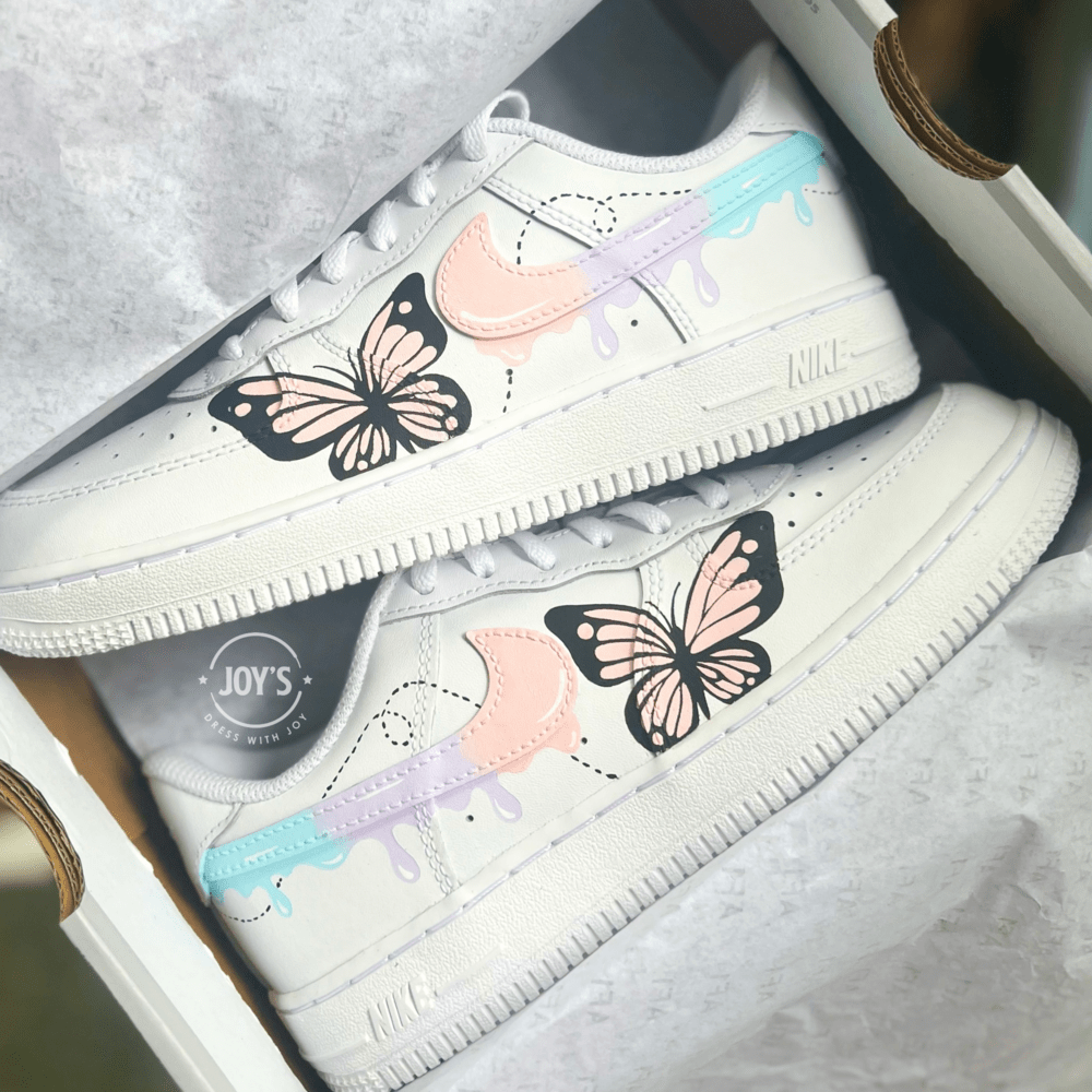 Custom white Nike Air Force 1 sneakers in a shoebox, featuring hand-painted butterfly designs and pastel drip swooshes in peach, lavender, and light blue.