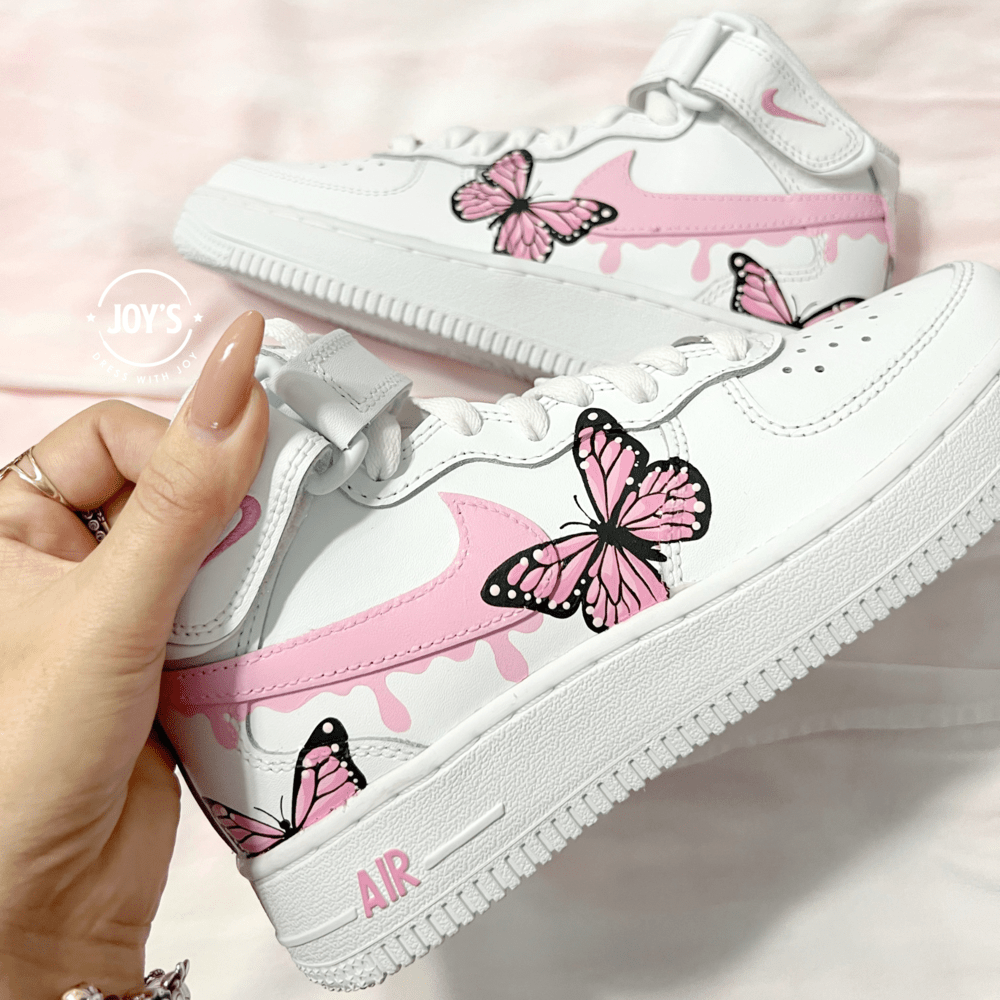 Pair of custom white Nike Air Force 1 sneakers with pink drip swoosh and hand-painted pink butterfly designs, displayed side by side on a soft pink background.