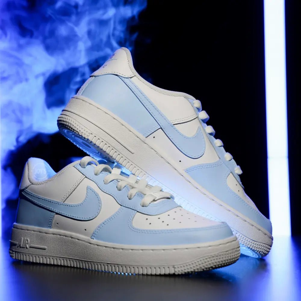 White and baby blue Air Force 1 sneakers photographed with blue smoke lighting on a dark background.