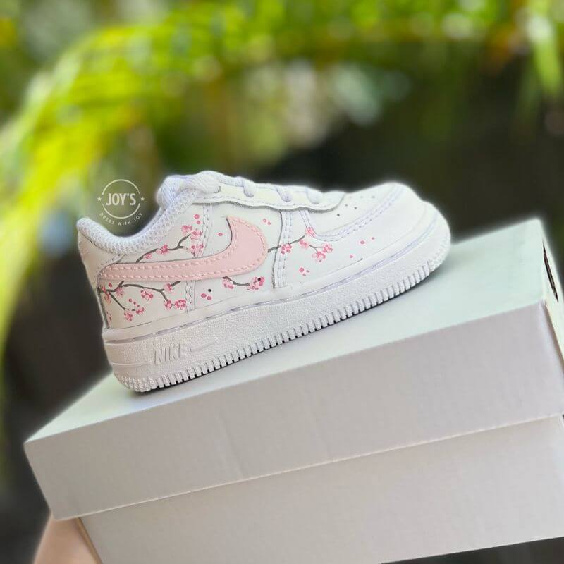 White Nike Air Force 1 toddler sneakers with soft pink swoosh and cherry blossom branch details, shown from the side.
