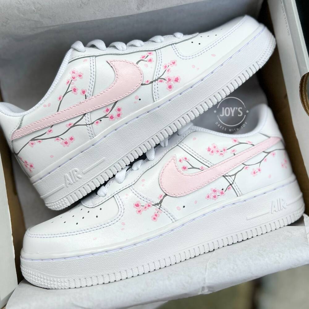 White Nike Air Force 1 with soft pink swoosh and hand-painted cherry blossom branches, shown in close-up.
