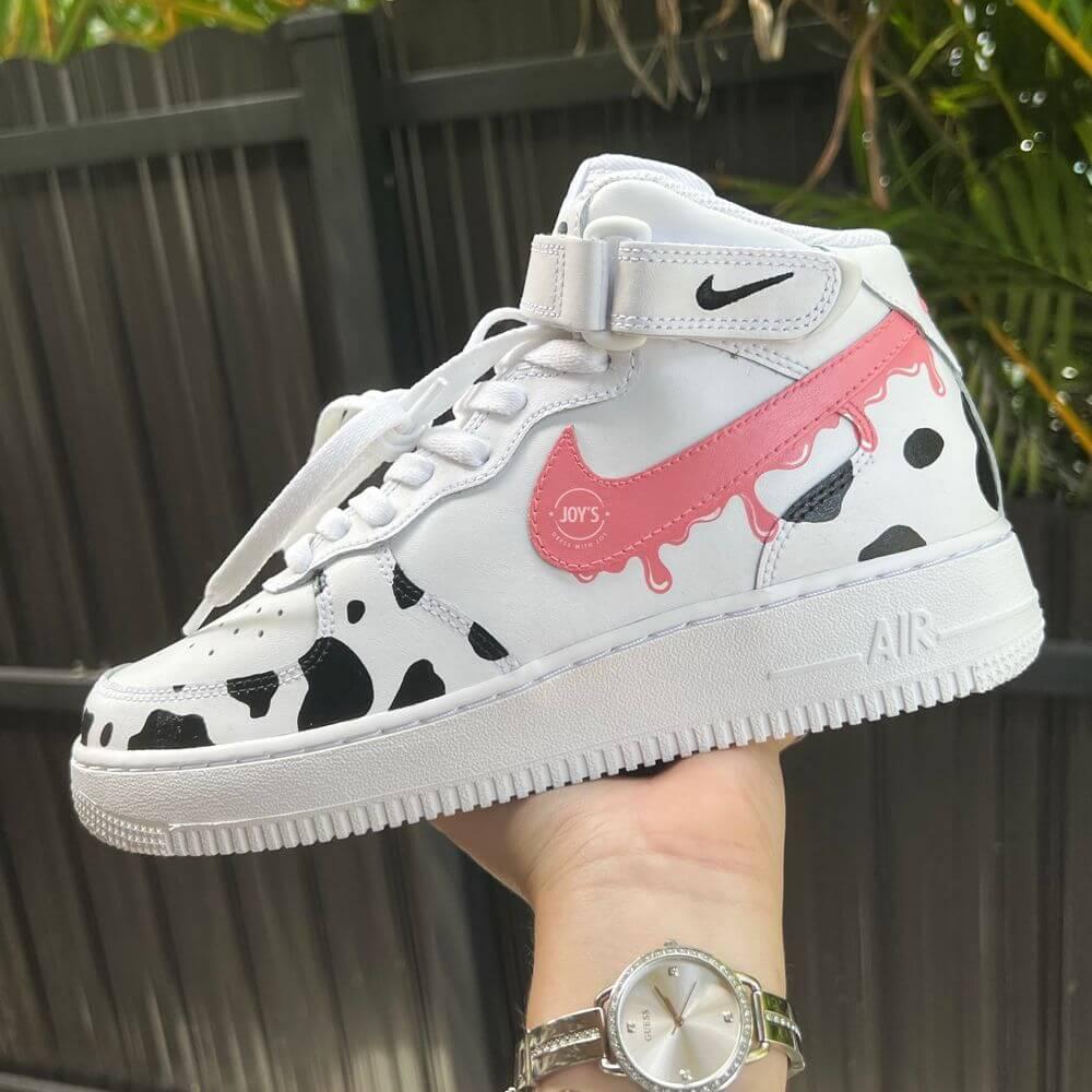 White Nike Air Force 1 mid-top sneaker with black cow print panels and a pink dripping swoosh, held in hand outdoors.