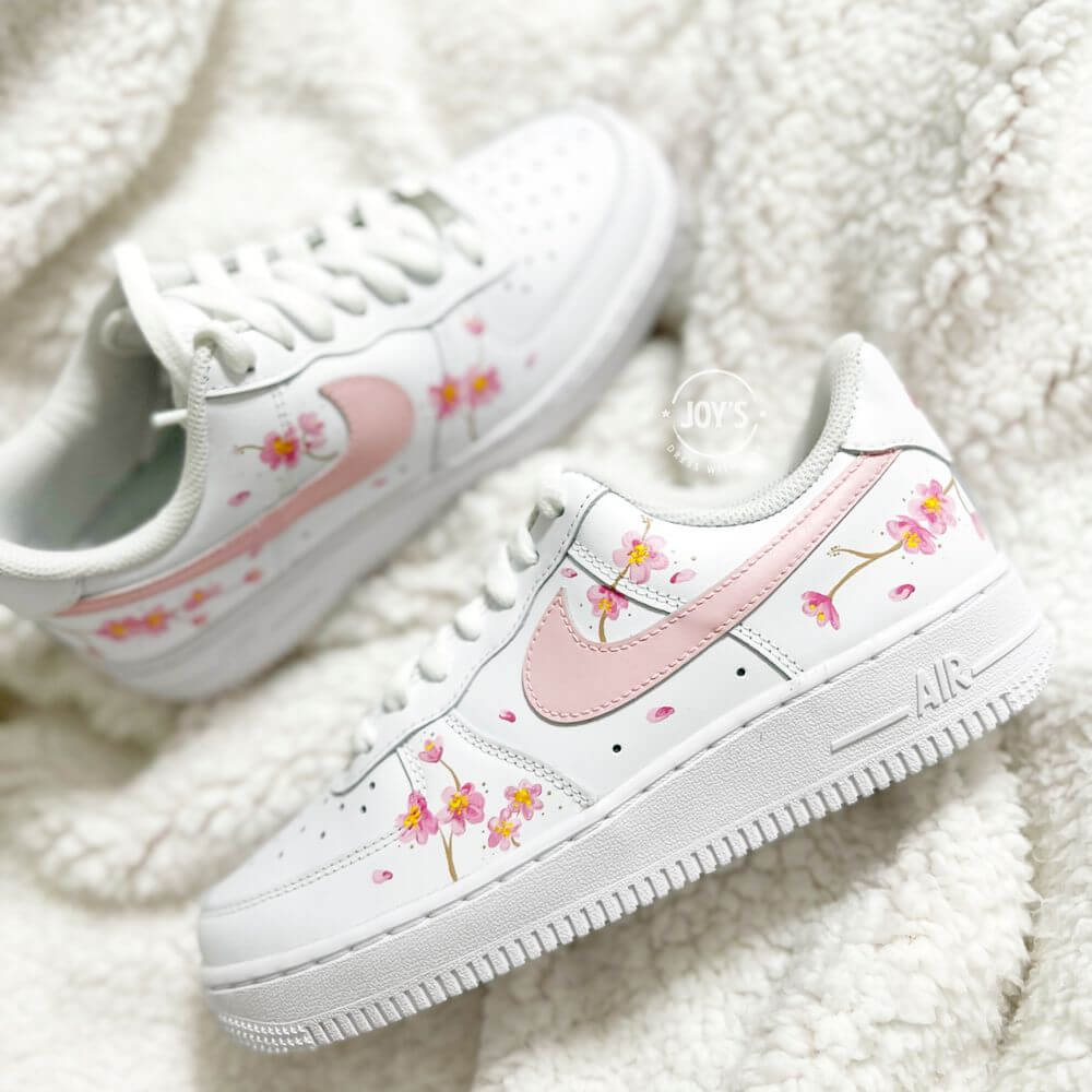 White Nike Air Force 1 sneaker with hand-painted pink cherry blossom flowers and a soft pink swoosh, shown close-up on a plush white background.