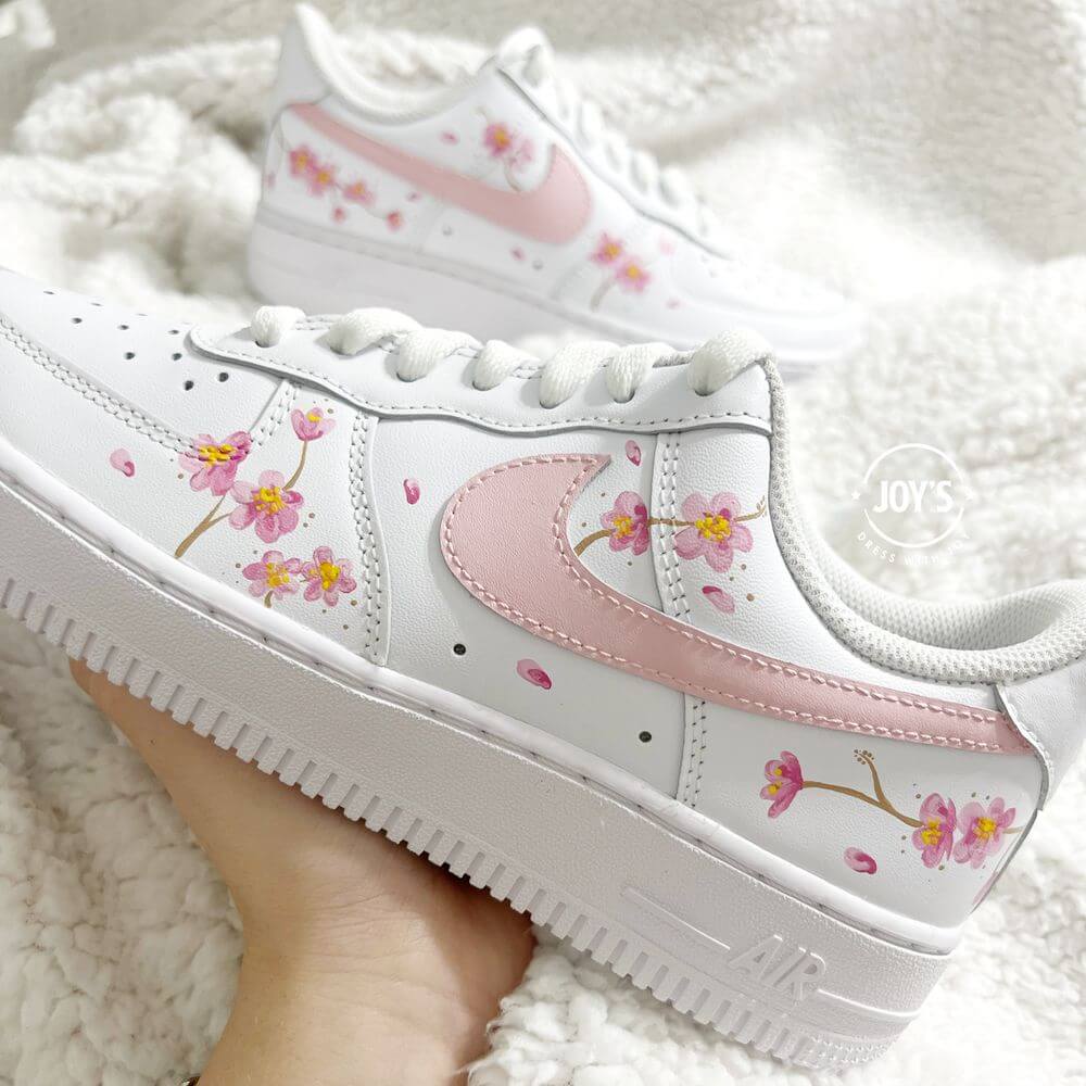 White Nike Air Force 1 sneaker with hand-painted pink cherry blossom flowers and a soft pink swoosh, shown close-up on a plush white background.