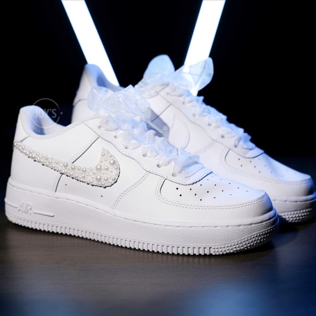 Custom white wedding Nike Air Force 1 with pearl-embellished swoosh, white ribbon laces, and personalized initials “J & E” on the heel, shown held in hand with soft bokeh lights in the background.