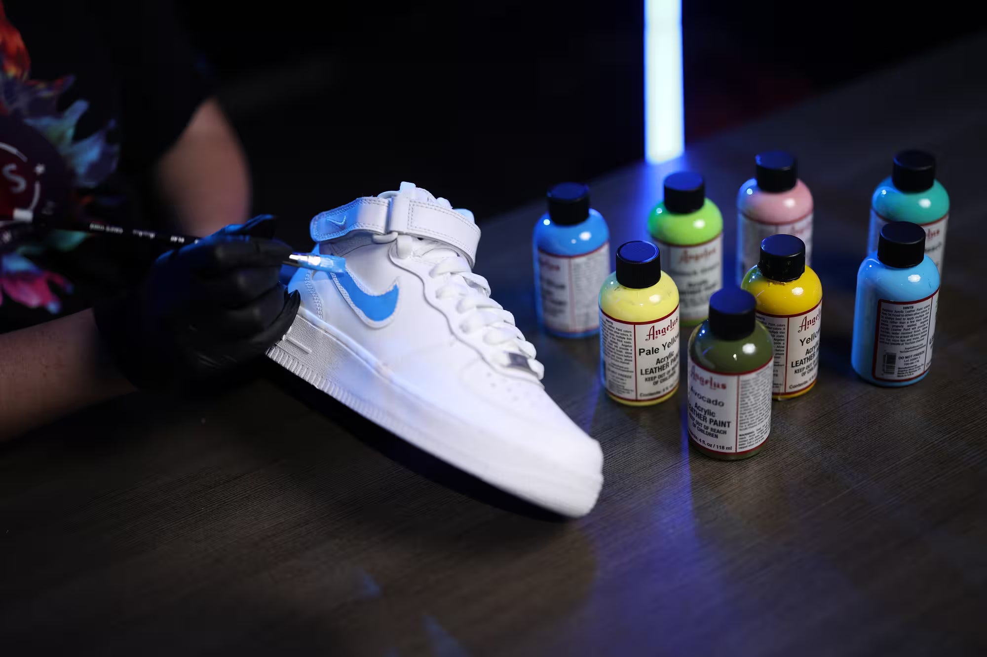 Joy Perez working on a white sneaker with glowing blue design, surrounded by colorful bottles of paint.