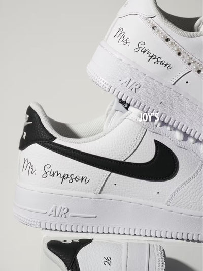 White custom wedding sneakers with pearl and rhinestone embellished swoosh, satin ribbon laces, and personalized last name printed on the side, shown in a close-up detail view