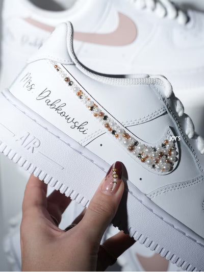 Custom white Nike Air Force 1 sneakers with subtle script lettering, one blush pink swoosh, and one swoosh embellished with pearls and rhinestones.
