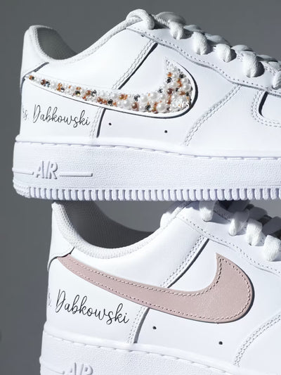 Custom white Nike Air Force 1 sneakers with subtle script lettering, one blush pink swoosh, and one swoosh embellished with pearls and rhinestones.
