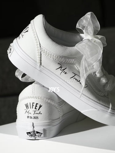 White custom wedding sneakers with pearl trim and personalized bride and groom names, wedding date, and “Wifey” and “Husband” heel text shown in a close-up detail view