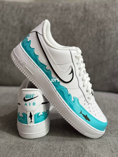 White custom Nike Air Force 1 sneakers with teal sea wave inspired hand-painted details and black swoosh with a shark figure