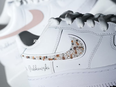 Custom white Nike Air Force 1 sneakers with subtle script lettering, one blush pink swoosh, and one swoosh embellished with pearls and rhinestones.
