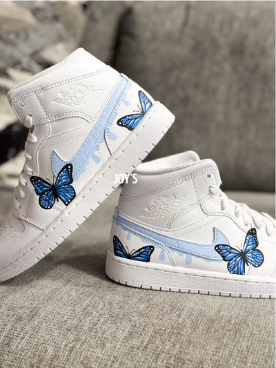 White custom Air Jordan 1 sneakers with hand-painted blue butterfly designs and light blue drip details on the sides, shown in a paired side view