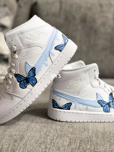 White custom Air Jordan 1 sneakers with hand-painted blue butterfly designs and light blue drip details on the sides, shown in a paired side view