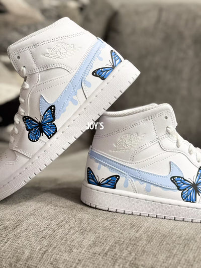 White custom Air Jordan 1 sneakers with hand-painted blue butterfly designs and light blue drip details on the sides, shown in a paired side view