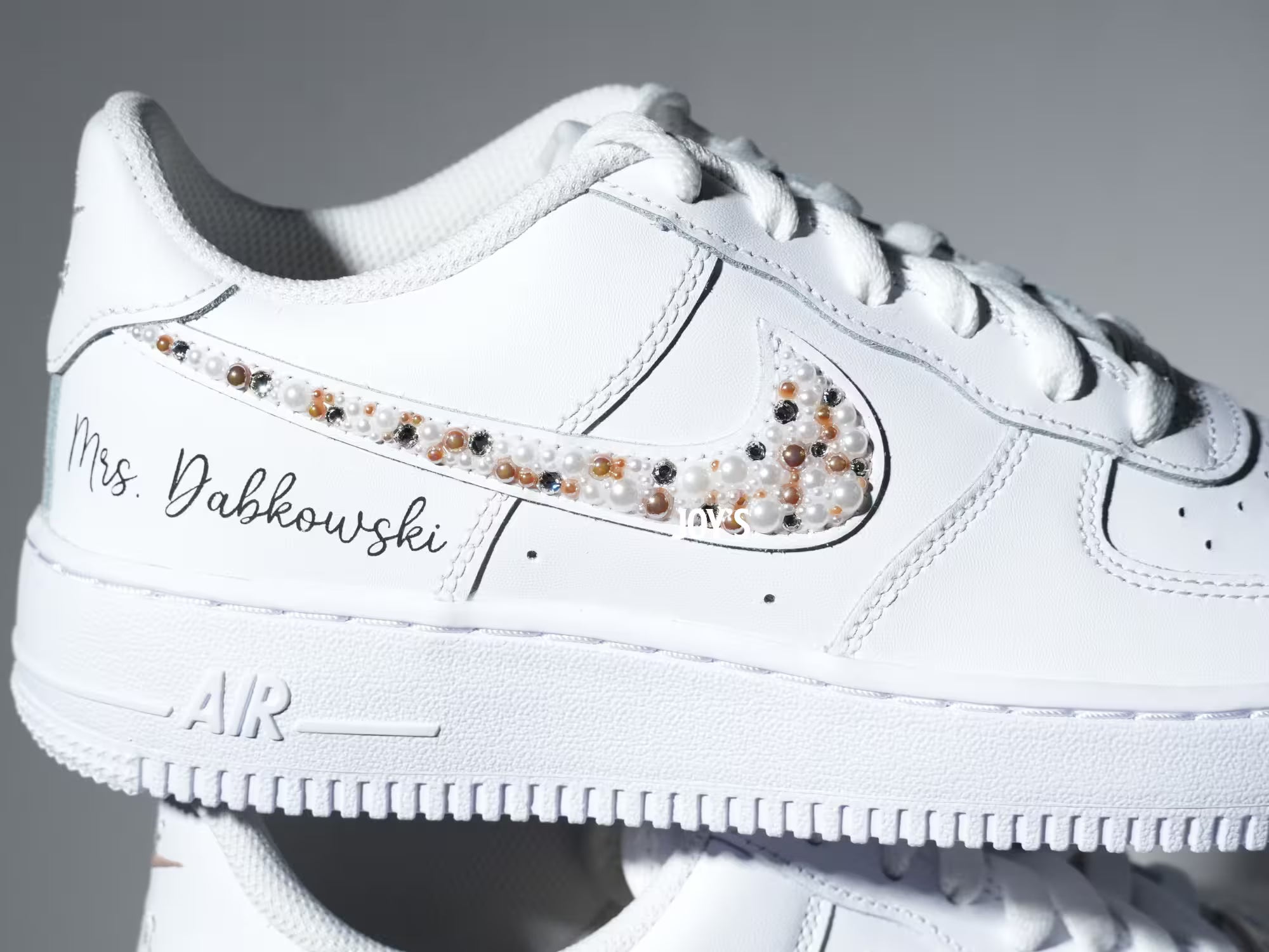 Custom white Nike Air Force 1 sneakers with subtle script lettering, one blush pink swoosh, and one swoosh embellished with pearls and rhinestones.
