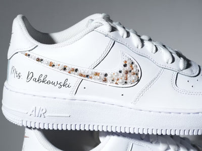 Custom white Nike Air Force 1 sneakers with subtle script lettering, one blush pink swoosh, and one swoosh embellished with pearls and rhinestones.
