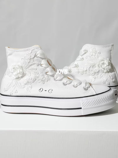 White platform Converse Chuck Taylor wedding sneakers with lace floral appliqués, satin ribbon laces, and personalized initials printed on the side, shown in a clean studio side view
