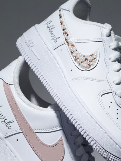Custom white Nike Air Force 1 sneakers with subtle script lettering, one blush pink swoosh, and one swoosh embellished with pearls and rhinestones.
