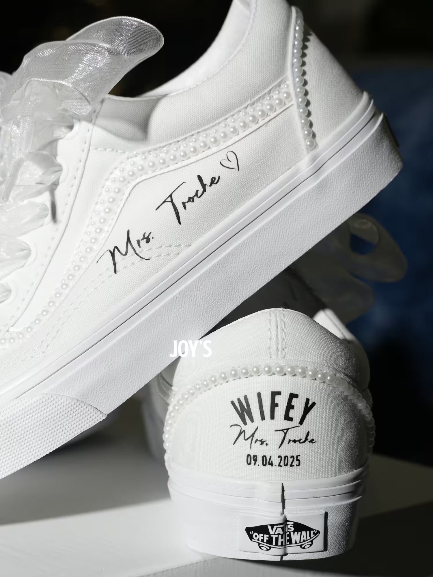 White custom wedding sneakers with pearl trim and personalized bride name, wedding date, and “Wifey” heel text shown in a close-up detail view