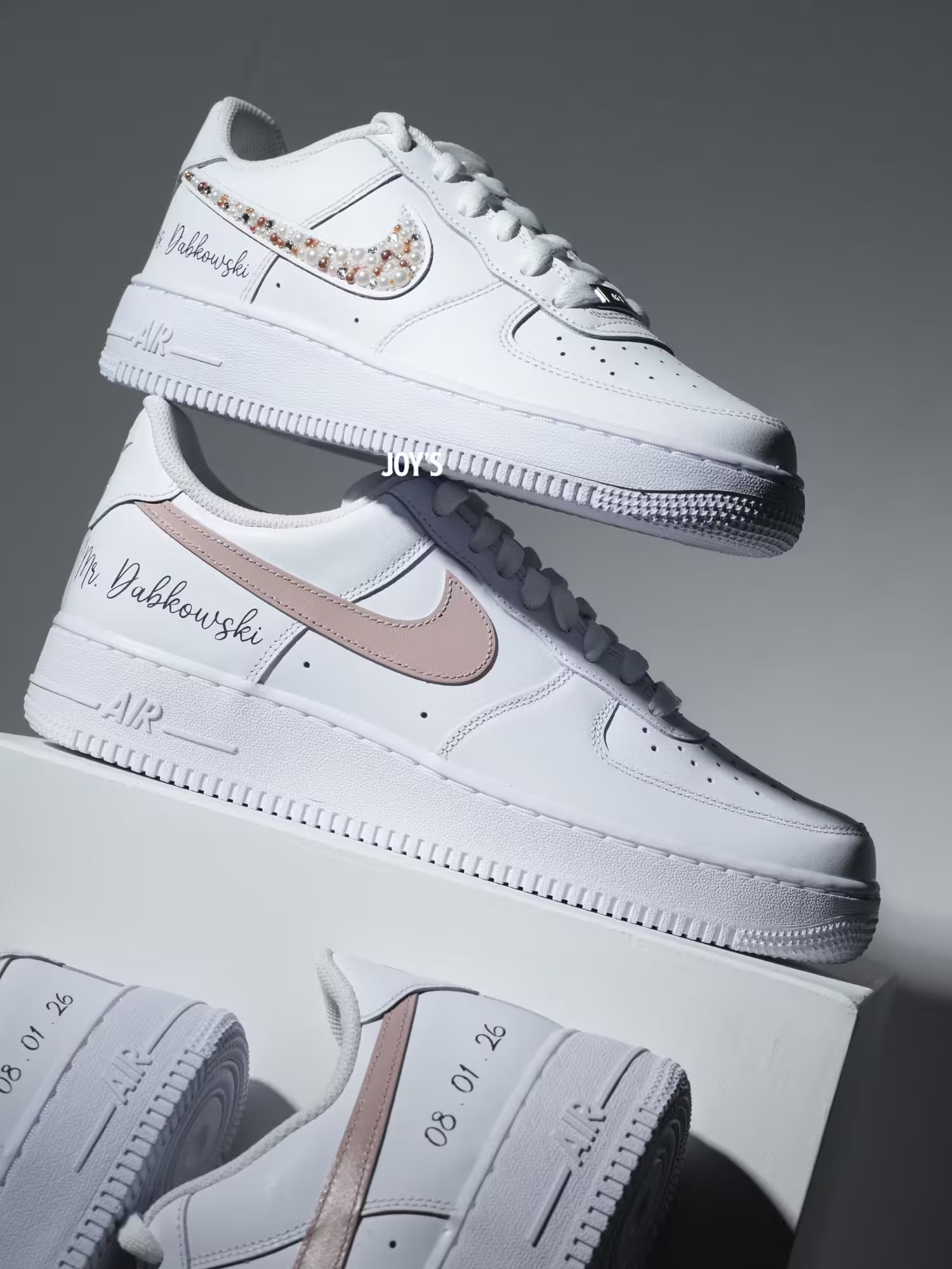 Custom white Nike Air Force 1 sneakers with subtle script lettering, one blush pink swoosh, and one swoosh embellished with pearls and rhinestones.
