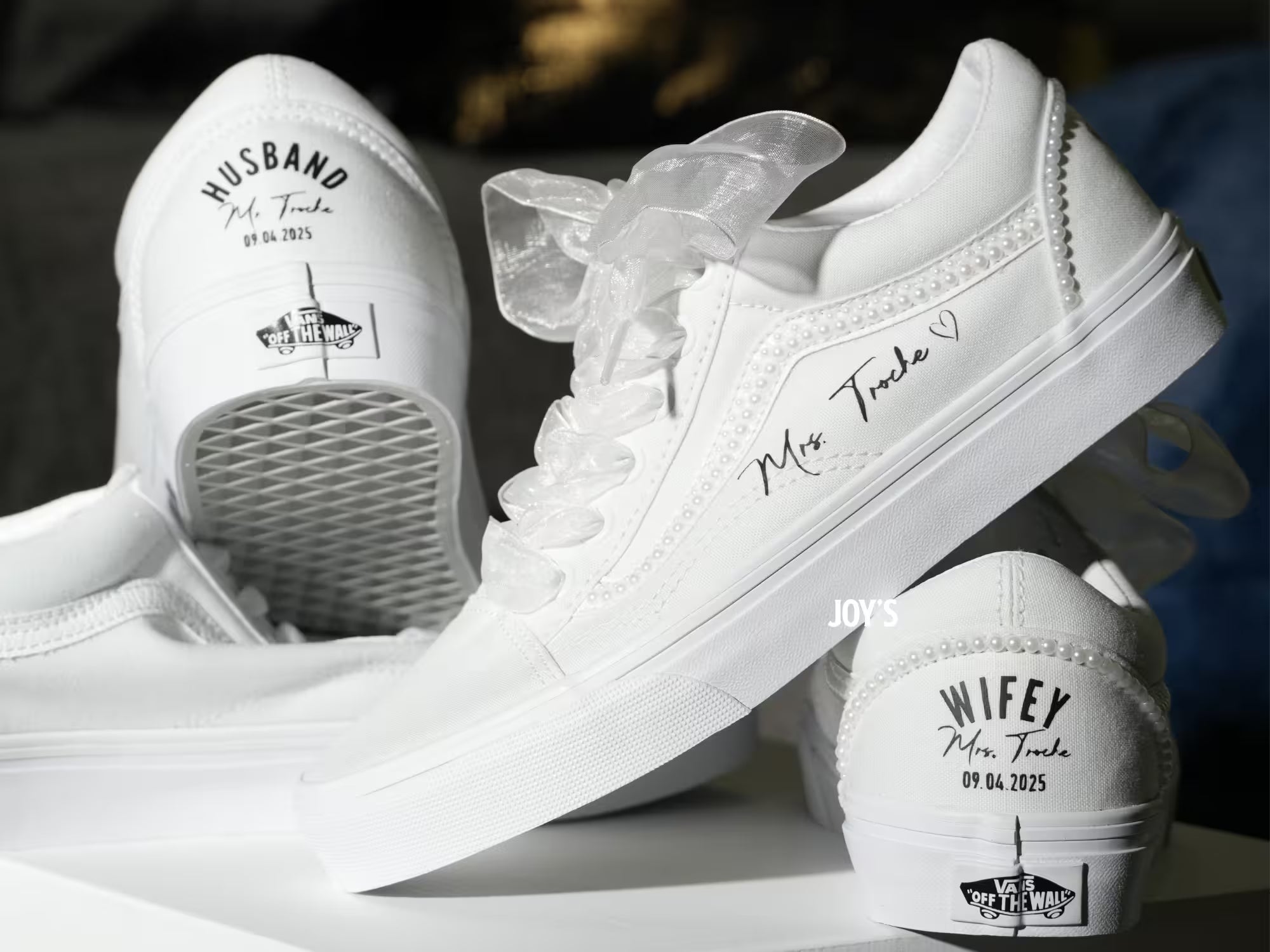 White custom wedding sneakers with pearl trim and personalized bride and groom names, wedding date, and “Wifey” and “Husband” heel text shown in a close-up detail view