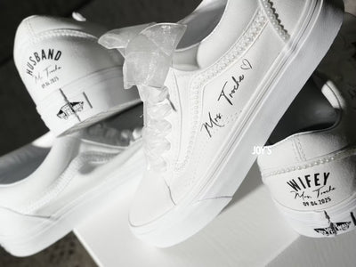 White custom wedding sneakers with pearl trim and personalized bride and groom names, wedding date, and “Wifey” and “Husband” heel text shown in a close-up detail view