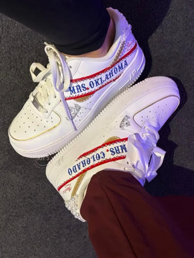 White custom wedding sneakers with pearl-embellished heel, rhinestone details, red and blue painted accents, and personalized “Mrs. Oklahoma” text on the side, shown in a close-up side view
