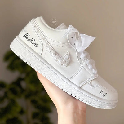 White Nike Air Force 1 Low sneakers (standard sole, not platform), customized with a crystal-embellished swoosh and subtle rhinestone detailing along the panels. Finished with sheer white ribbon laces and personalized black script on the heel and toe, giving a clean, elegant bridal look.
