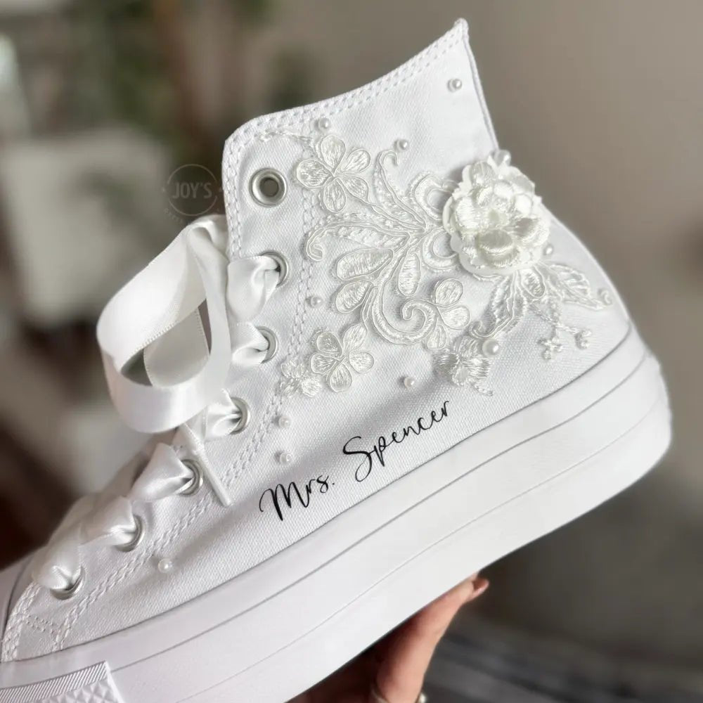 White platform Vans wedding sneakers with lace floral appliqués, satin ribbon laces, and personalized bride name printed on the side, shown in a close-up angled view