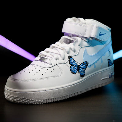 White Nike Air Force 1 Mid Sneakers with light blue drip swoosh and hand-painted blue butterfly design.