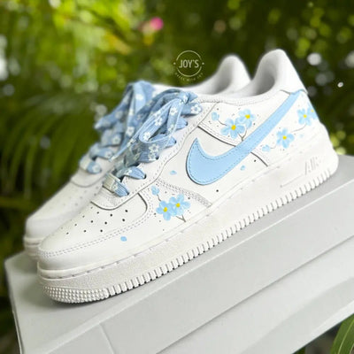 White Nike Air Force 1 with light blue swoosh, blue floral accents, and matching blue patterned laces, displayed on a white box.