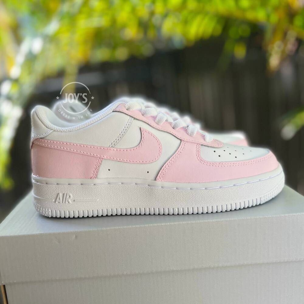White Nike Air Force 1 Low-top sneakers with soft pink panels and swoosh, displayed on a white box with palm leaves in the background.