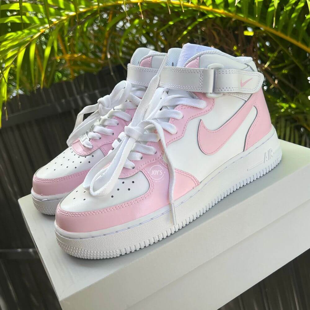 White Nike Air Force 1 Mid-top sneakers with soft pink panels and swoosh, displayed on a white box with palm leaves in the background.