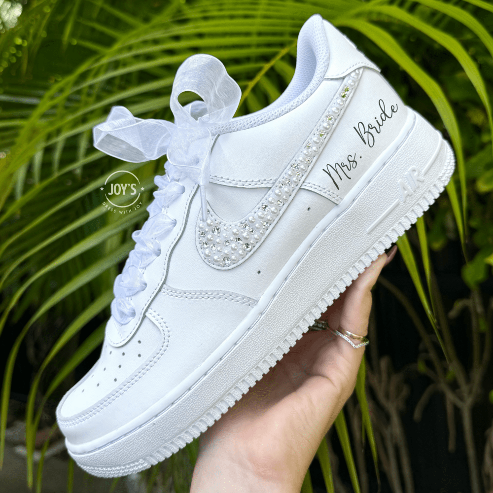 Custom wedding white Nike Air Force 1 with pearl and rhinestone swoosh, white ribbon laces, and the name Mrs. personalized on the heel, shown held in hand with soft holiday lights in the background.